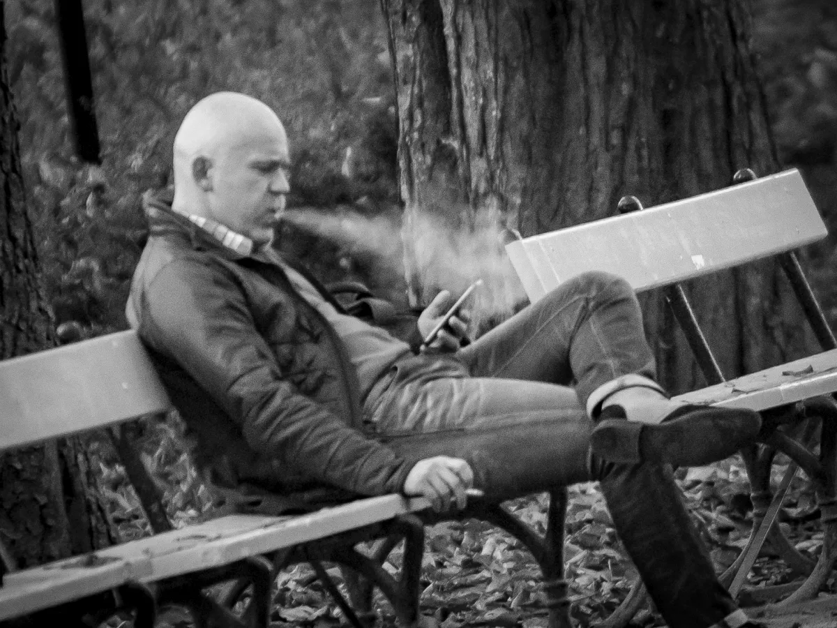 Bald man smoking on a bench while looking down at his phone